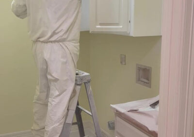 cabinet painting technician in protective suit spraying upper cabinets on ladder