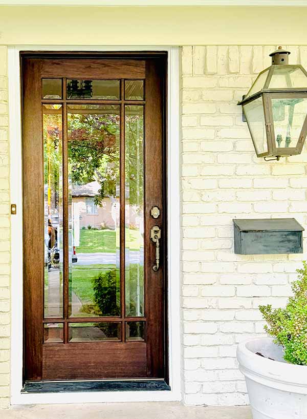 Stained wood front door with glass panels next to painted white brick and gas lantern in North Dallas