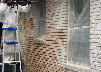 technician in protective suit pressure washing paint from exterior brick wall on ladder