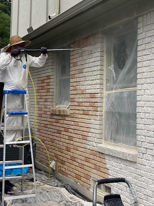 technician in protective suit pressure washing paint from exterior brick wall on ladder