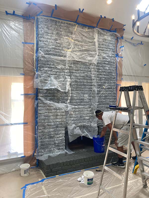 painter preparing interior brick fireplace for paint removal with plastic sheeting and blue tape