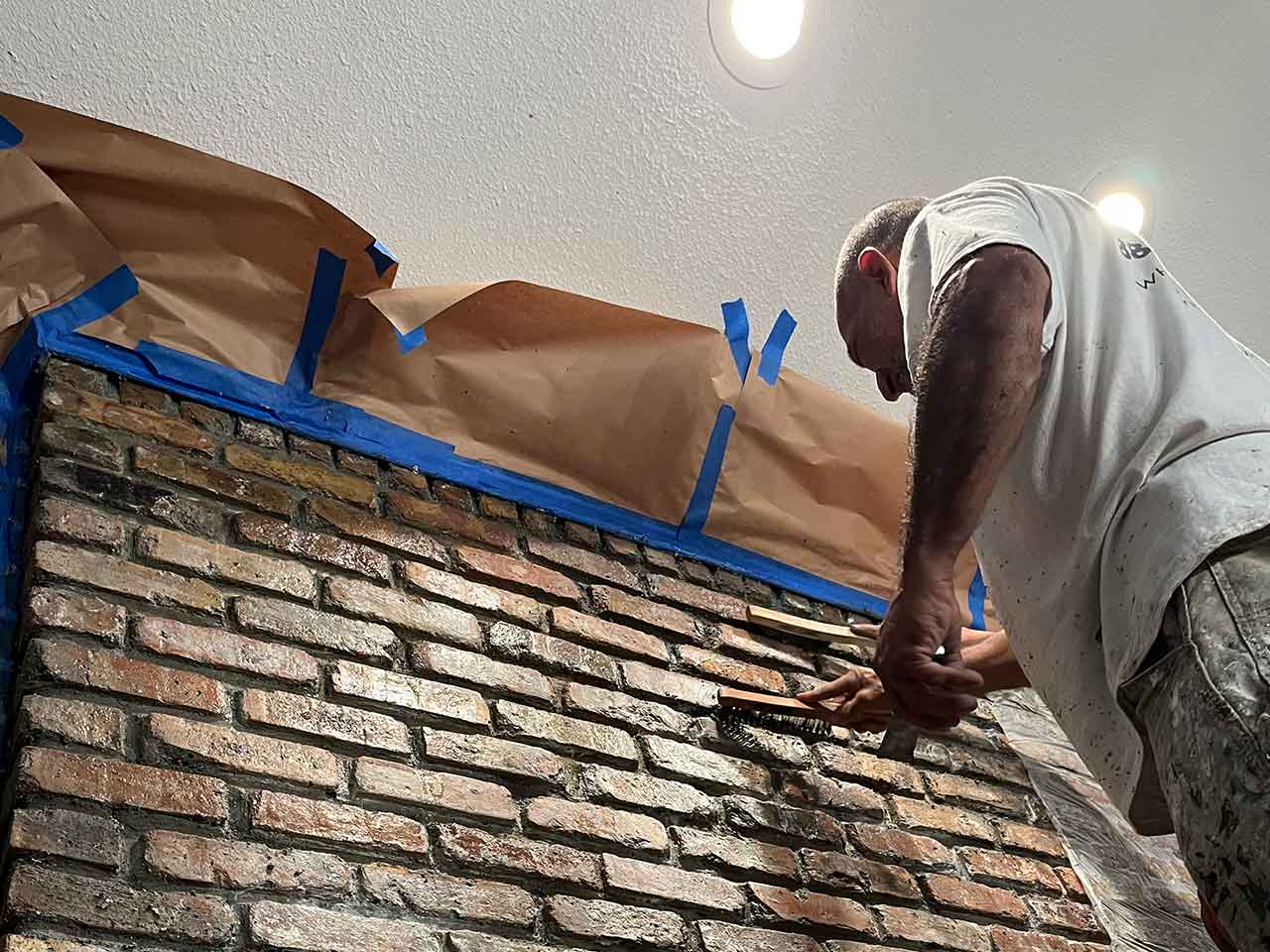 Interior Textured Brick Paint Stripping — PPP North Dallas Technician using a wire brush to strip paint from textured brick indoors with protective masking on ceiling and adjacent wall